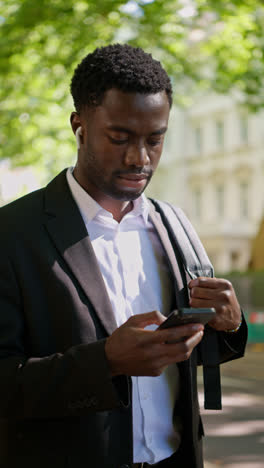 Vertical-Video-Of-Young-Businessman-Wearing-Wireless-Earbuds-Streaming-Music-Or-Podcast-Checking-Mobile-Phone-On-Walk-To-Work-In-Offices-In-The-Financial-District-Of-The-City-Of-London-UK---shot-in-real-time